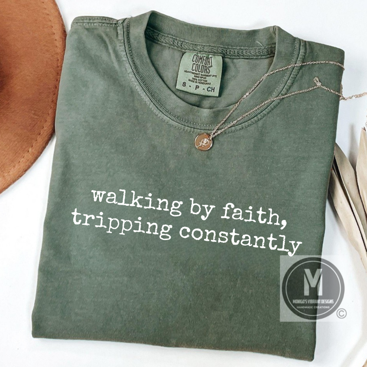 Folded green t-shirt with text 'walking by faith, tripping constantly' on a white background