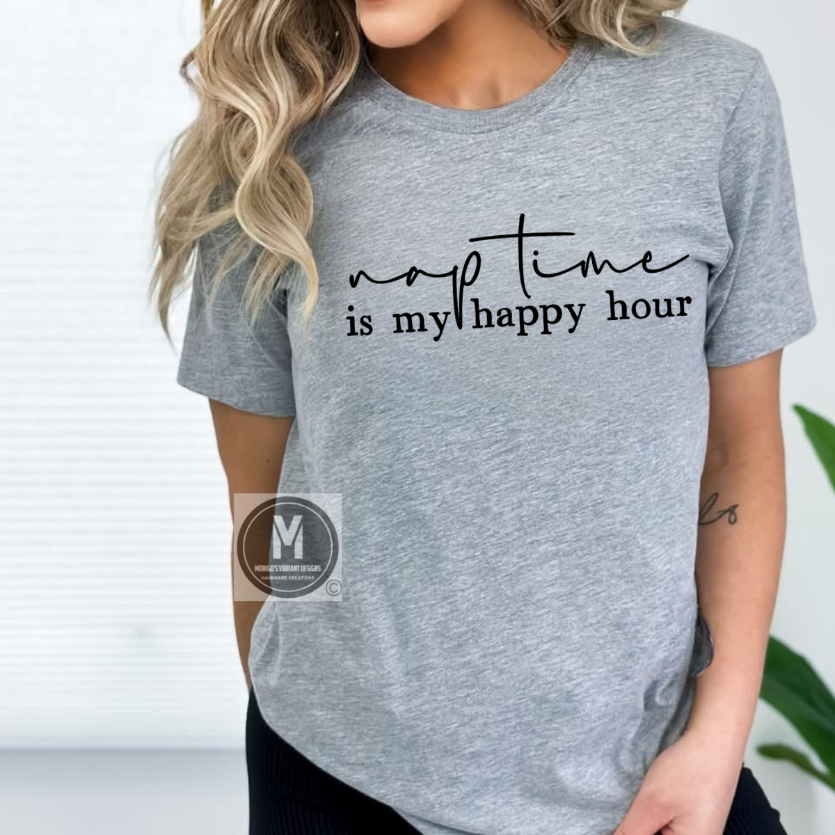 A woman in a gray t-shirt with "nap time is my happy hour" printed in black script, smiling against a light window backdrop.