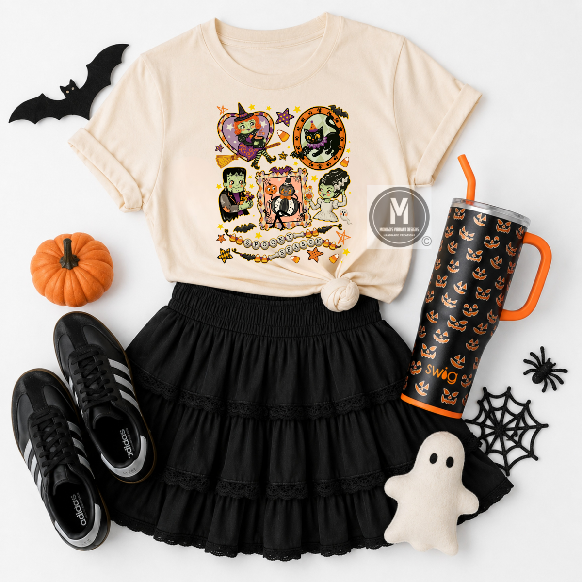A styled flat-lay of a cream graphic T-shirt with a Halloween-themed collage print, paired with a black ruffled skirt, black sneakers, a black tumbler with orange accents, a small ghost plush, and autumn props on a white background.