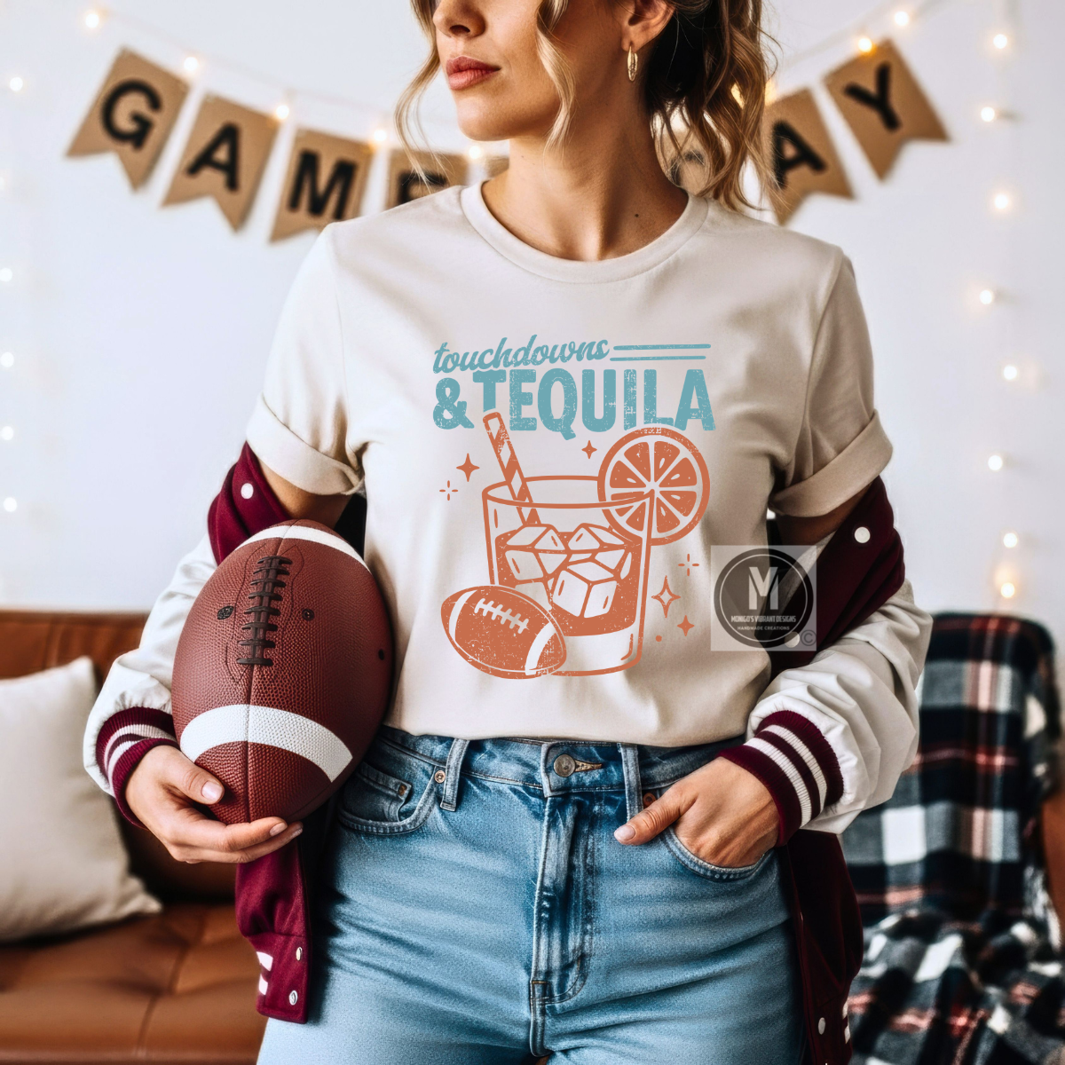 A woman wearing a cream graphic t-shirt with a 'touchdowns & TEQUILA' design, styled with a maroon and white varsity jacket, holding a football in a cozy, game-day decorated room.