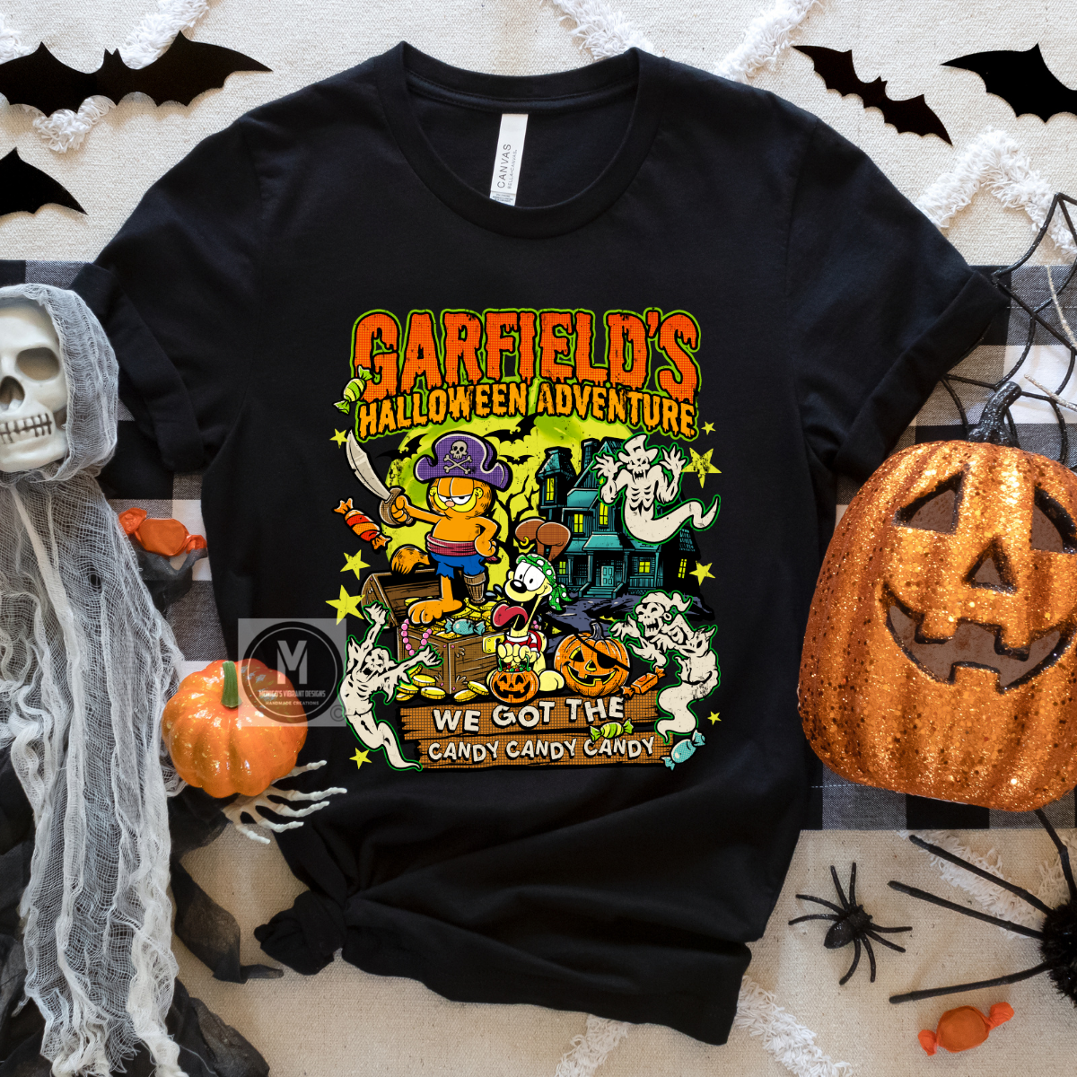 A black short-sleeve graphic T-shirt laid flat on a Halloween-themed backdrop with a sequined jack-o'-lantern, spiderwebs, a skull, and candy props. The shirt features a colorful cartoon-style front print reading 'Garfield's Halloween Adventure' and 'We Got the Candy Candy Candy' with Garfield, ghosts, a haunted house, and pumpkins.