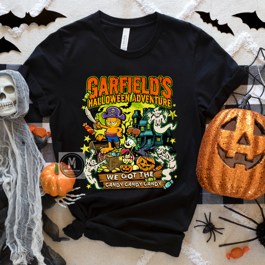A black short-sleeve graphic T-shirt laid flat on a Halloween-themed backdrop with a sequined jack-o'-lantern, spiderwebs, a skull, and candy props. The shirt features a colorful cartoon-style front print reading 'Garfield's Halloween Adventure' and 'We Got the Candy Candy Candy' with Garfield, ghosts, a haunted house, and pumpkins.