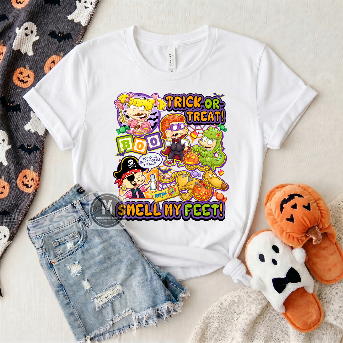 A white graphic t-shirt featuring a colorful cartoon Halloween design with the text 'TRICK OR TREAT! BOO SMELL MY FEET!', styled in a flat lay with denim shorts, a ghost-patterned blanket, and orange plush slippers.